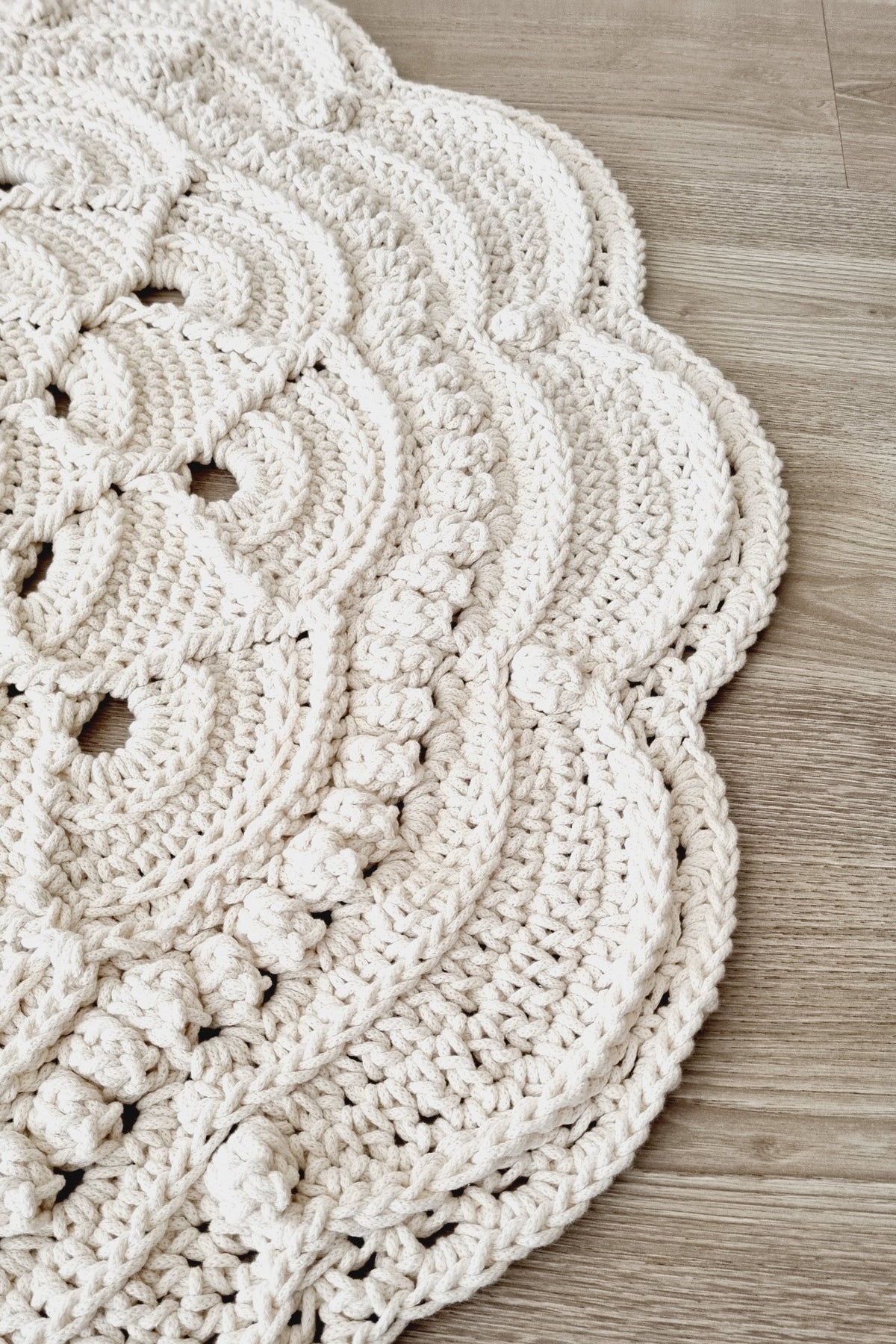 A close-up of a chunky crocheted rug in a warm off-white/cream color, featuring a scalloped edge and textured bobble stitch details, laid out on a light wood floor.