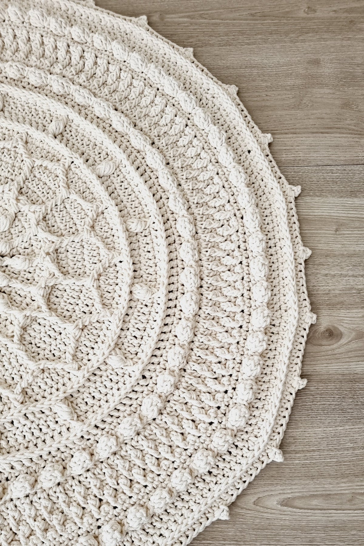 A round, hand-crocheted area rug in a warm cream or off-white color, featuring a textured bobble pattern, laid out on a light brown wooden floor.