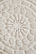 Close-up of a cream-colored crocheted textile with a detailed radial floral or mandala pattern, showing the texture and stitch details.
