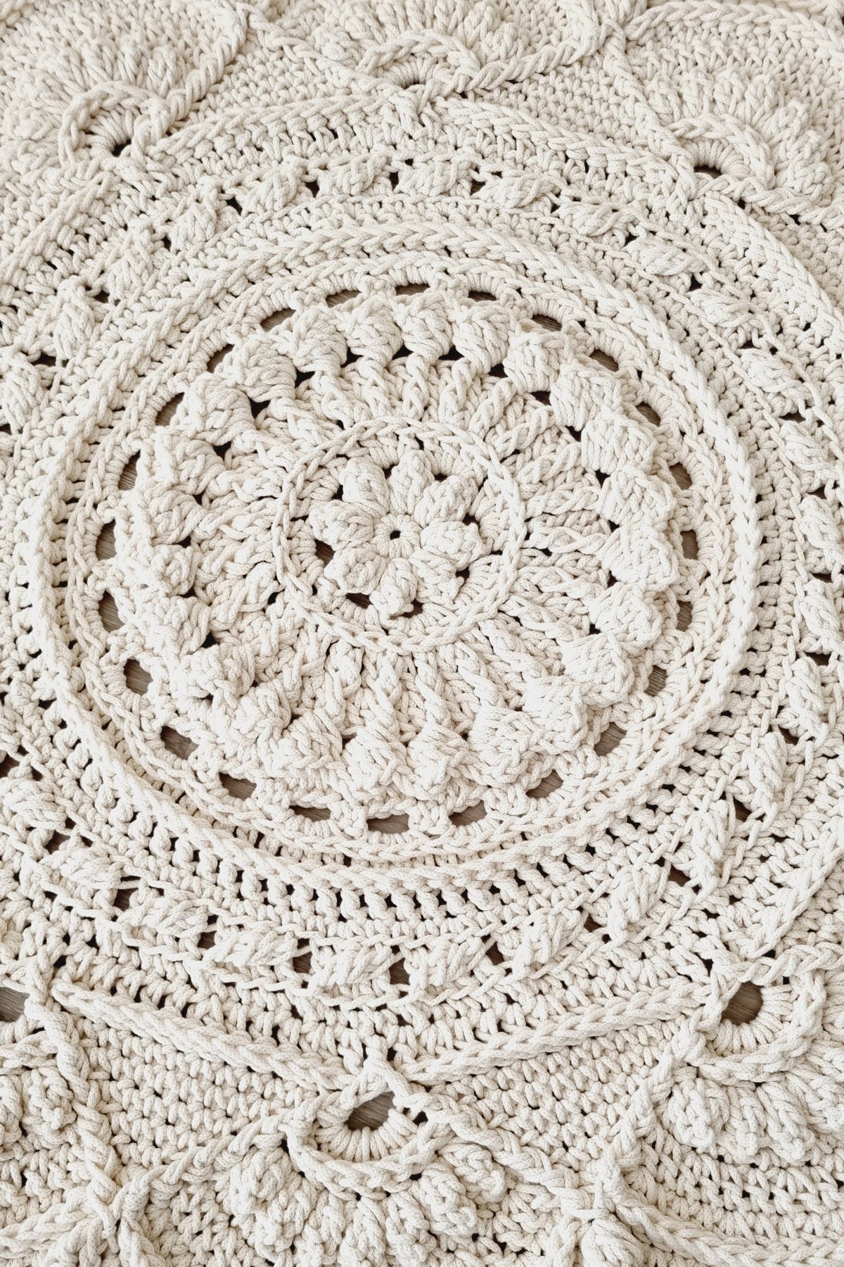 A close-up of a hand-crocheted round rug in a warm cream or off-white color, showcasing intricate circular and scalloped patterns on a light wood floor.