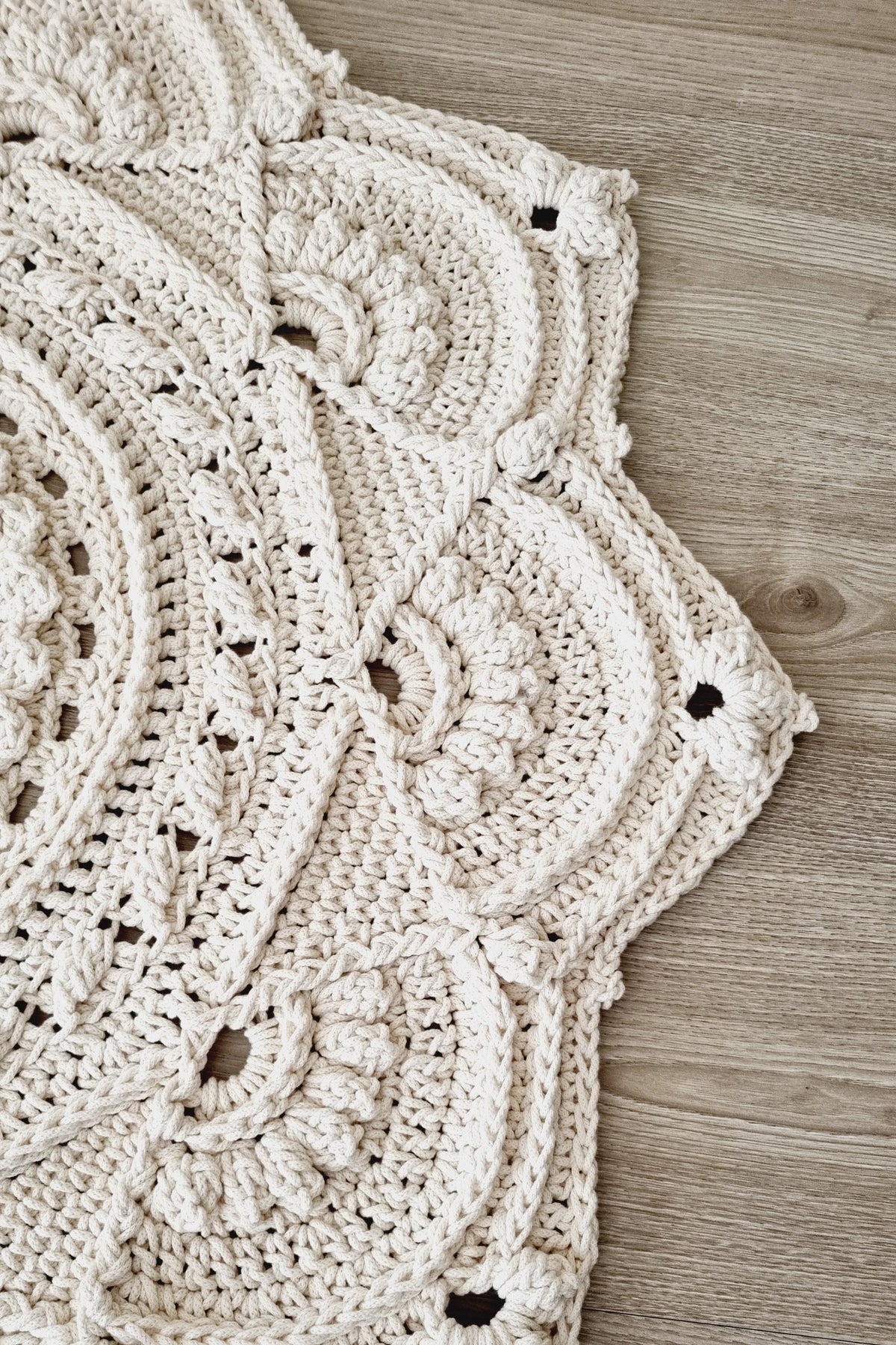 A close-up of a cream-colored crocheted floor rug with intricate floral and scalloped patterns, laid flat on a light wood floor.