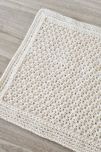 A close-up of a cream-colored crocheted floor rug laid flat on a light gray wood-grain floor.