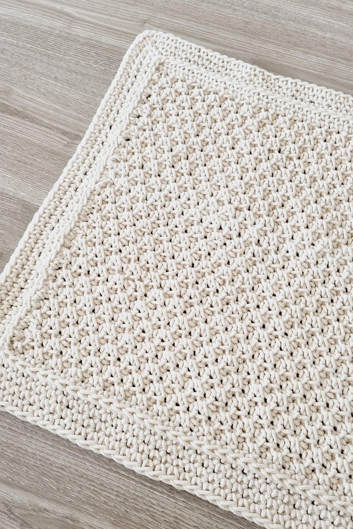 A close-up of a cream-colored crocheted floor rug laid flat on a light gray wood-grain floor.