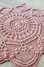 A dusty pink crocheted doily with intricate circular and scalloped patterns, laid flat on a light gray wood floor.