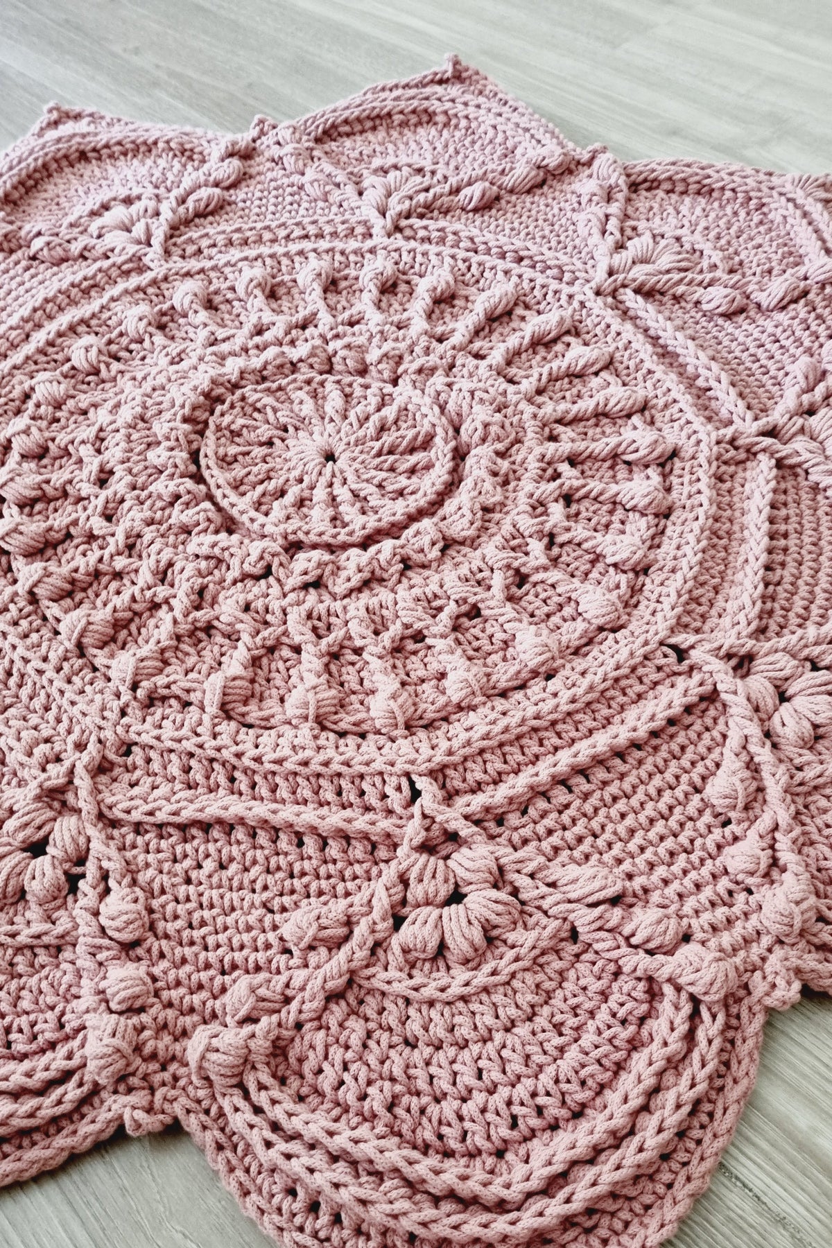 A dusty pink crocheted doily with intricate circular and scalloped patterns, laid flat on a light gray wood floor.
