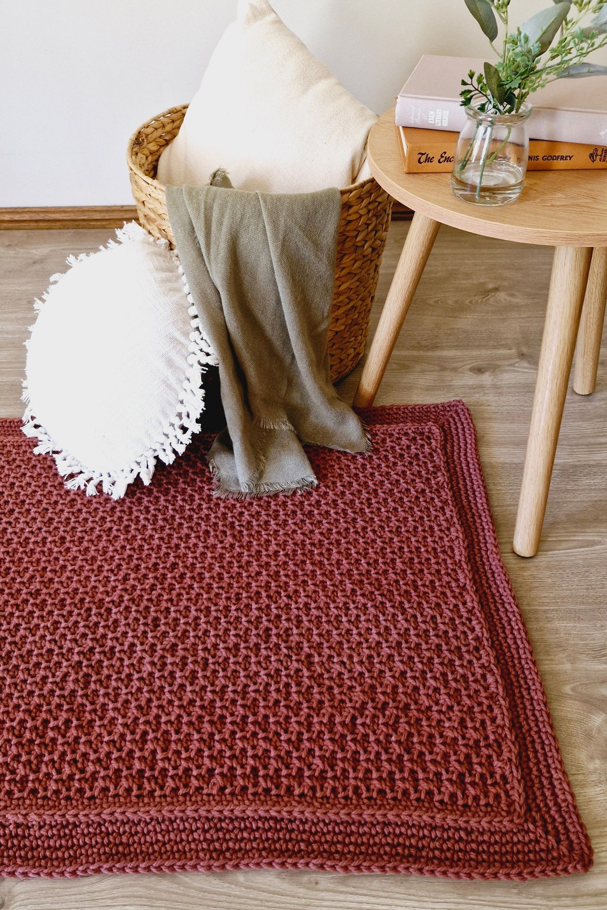 A textured chunky floor rug in a dusty terracotta red color, styled in a cozy room setting with a wooden side table, a woven basket, and decorative pillows.