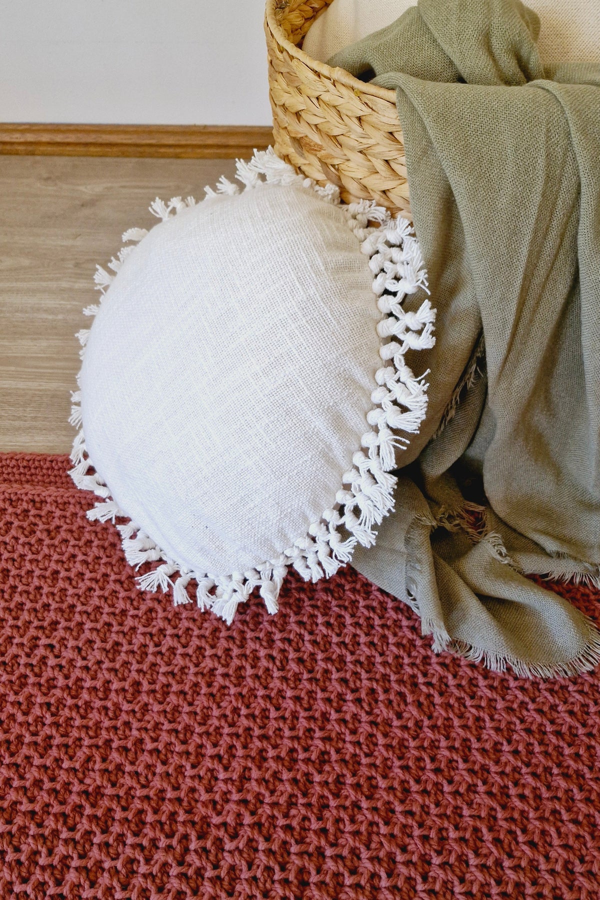 A round white textured throw pillow with white fringe trim, styled on a rust-red crochet floor rug next to a woven basket containing a sage green throw blanket.