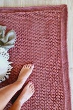 A brown hand-crocheted door rug with a textured pattern and a matching braided border, shown on a light wood floor with a person's bare feet and a white throw blanket.
