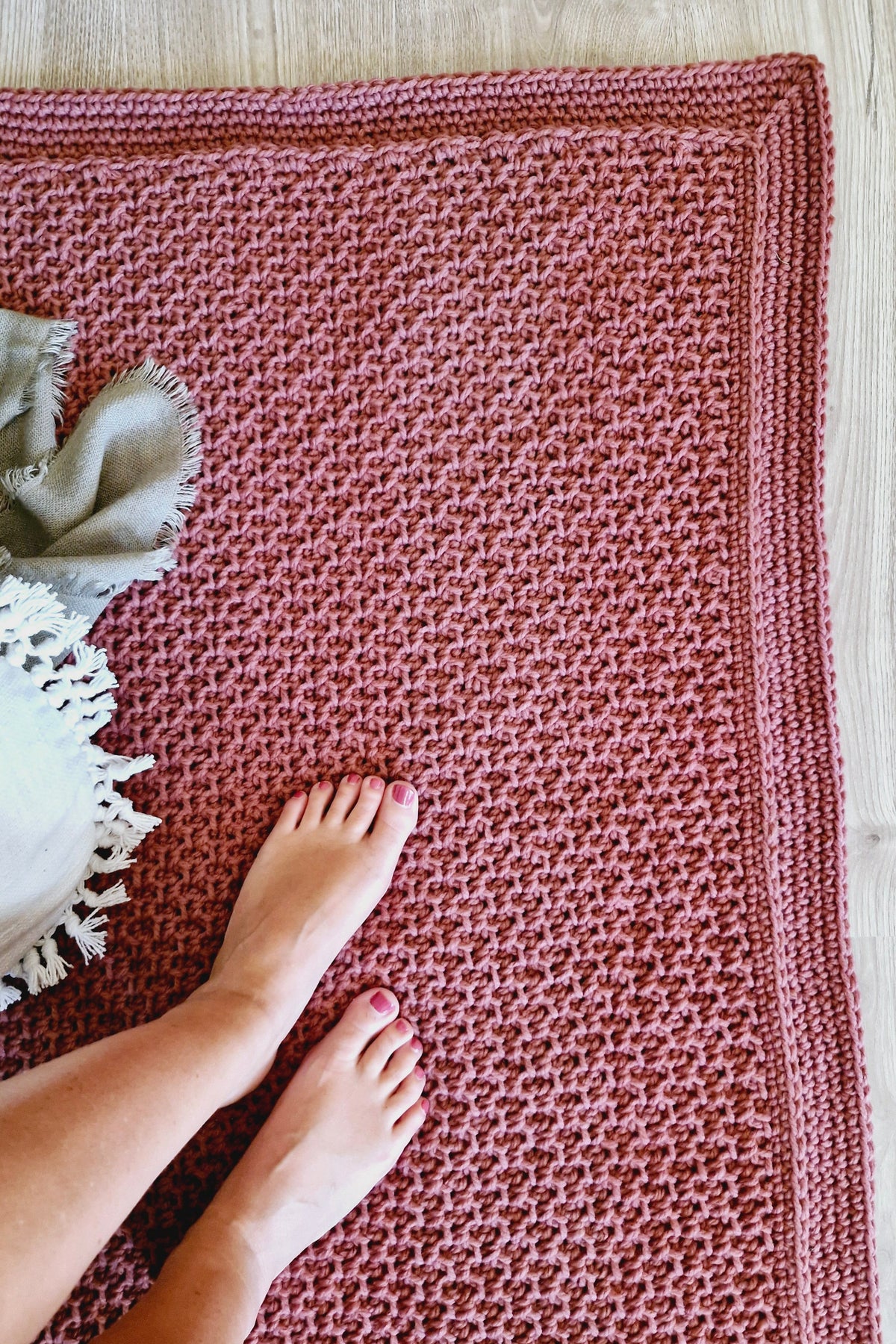 A brown hand-crocheted door rug with a textured pattern and a matching braided border, shown on a light wood floor with a person's bare feet and a white throw blanket.