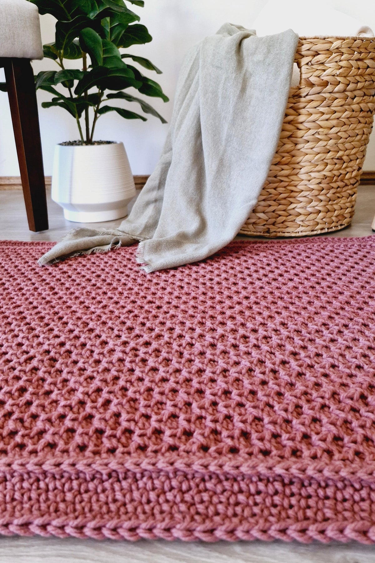 A brown crocheted floor rug with a chunky waffle texture, styled in a bright room with a potted green plant, a beige throw blanket, and a woven straw basket.
