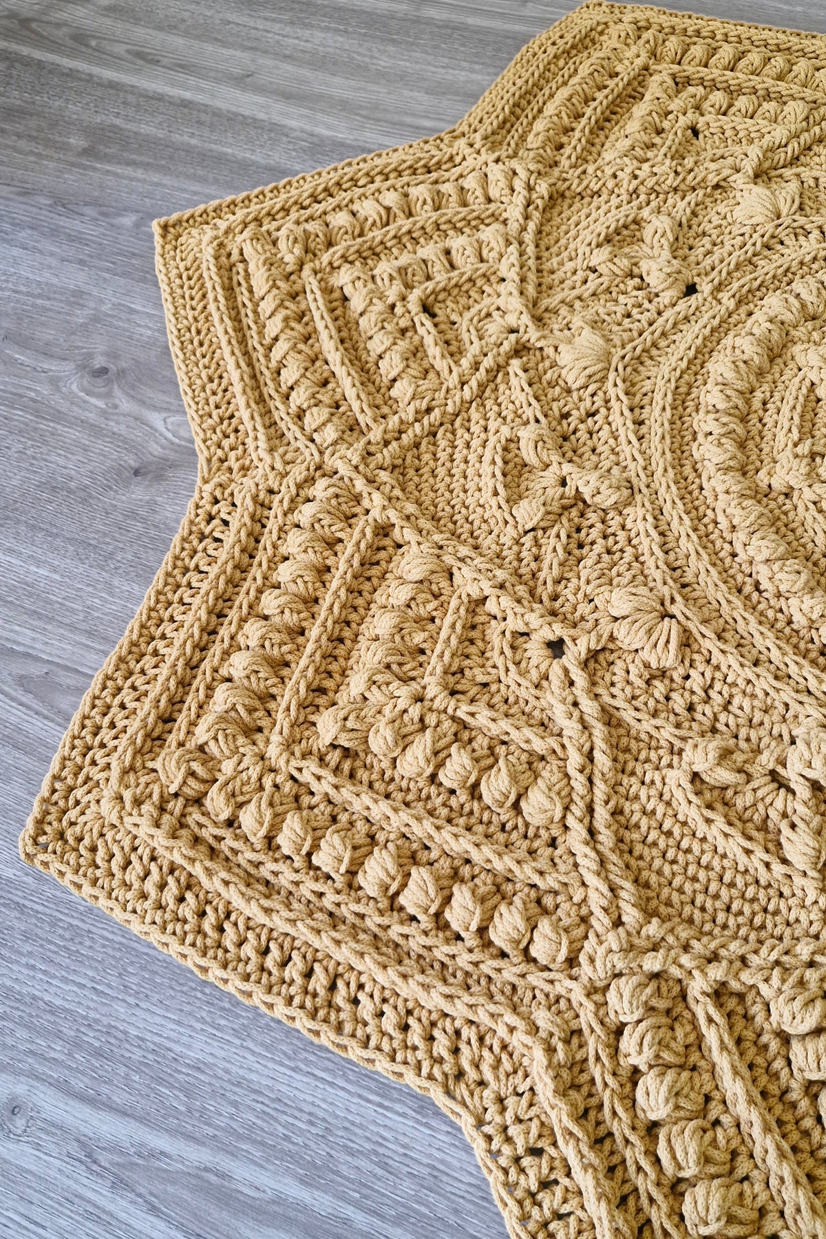 A chunky crocheted star-shaped rug in a warm mustard yellow color, laid out on a gray wood-plank floor.
