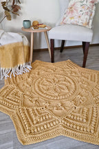 A large, star-shaped crocheted area rug in a warm mustard yellow color, styled in a cozy living room setting on a gray wood floor next to a light gray armchair and a small wooden side table.