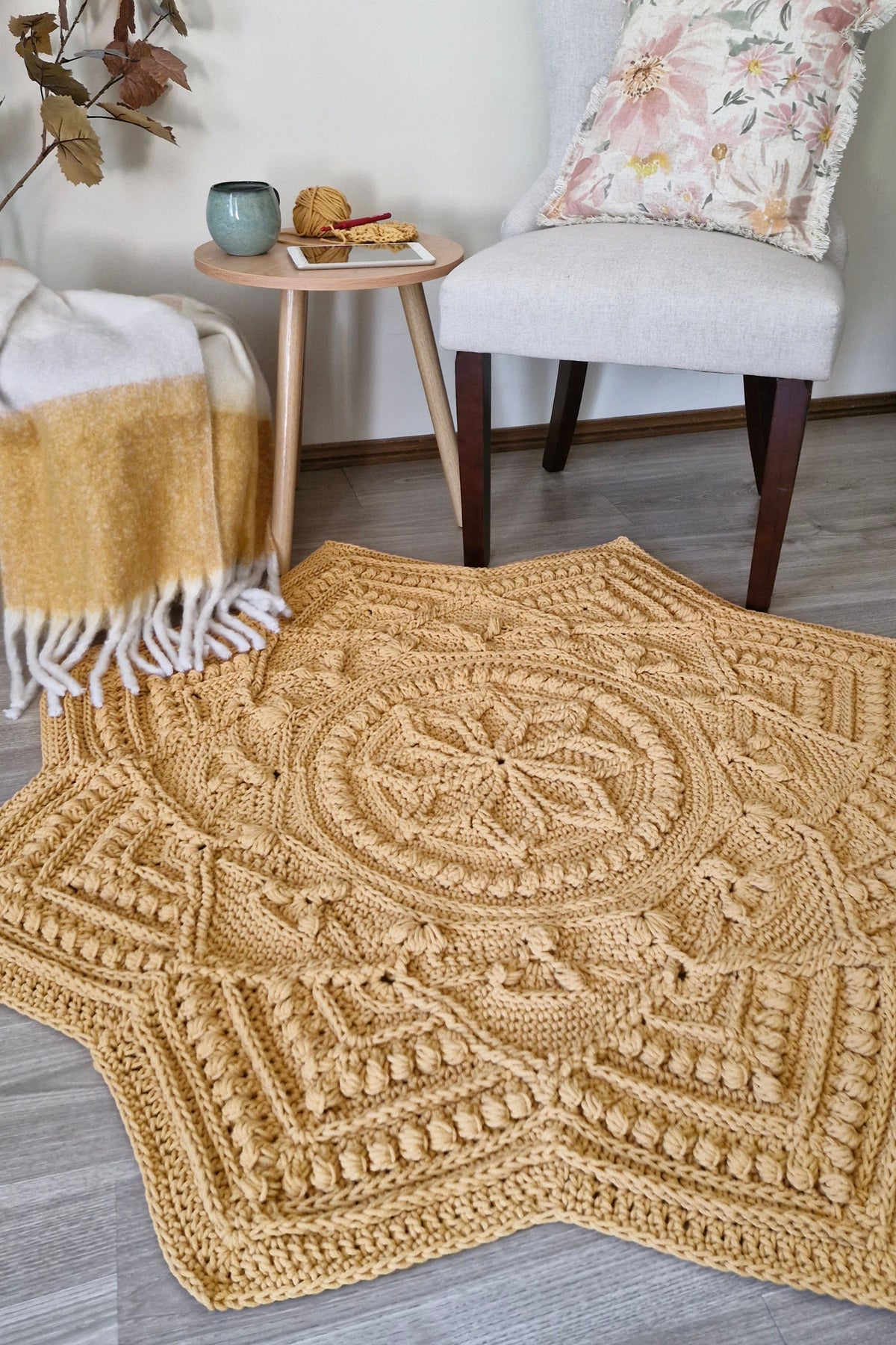 A large, star-shaped crocheted area rug in a warm mustard yellow color, styled in a cozy living room setting on a gray wood floor next to a light gray armchair and a small wooden side table.