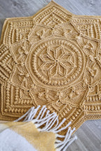 A large crocheted star-shaped crocher rug in a warm mustard-golden tan, laid flat on a gray wood floor. A white and mustard throw blanket with white fringe is partially visible in the lower foreground.