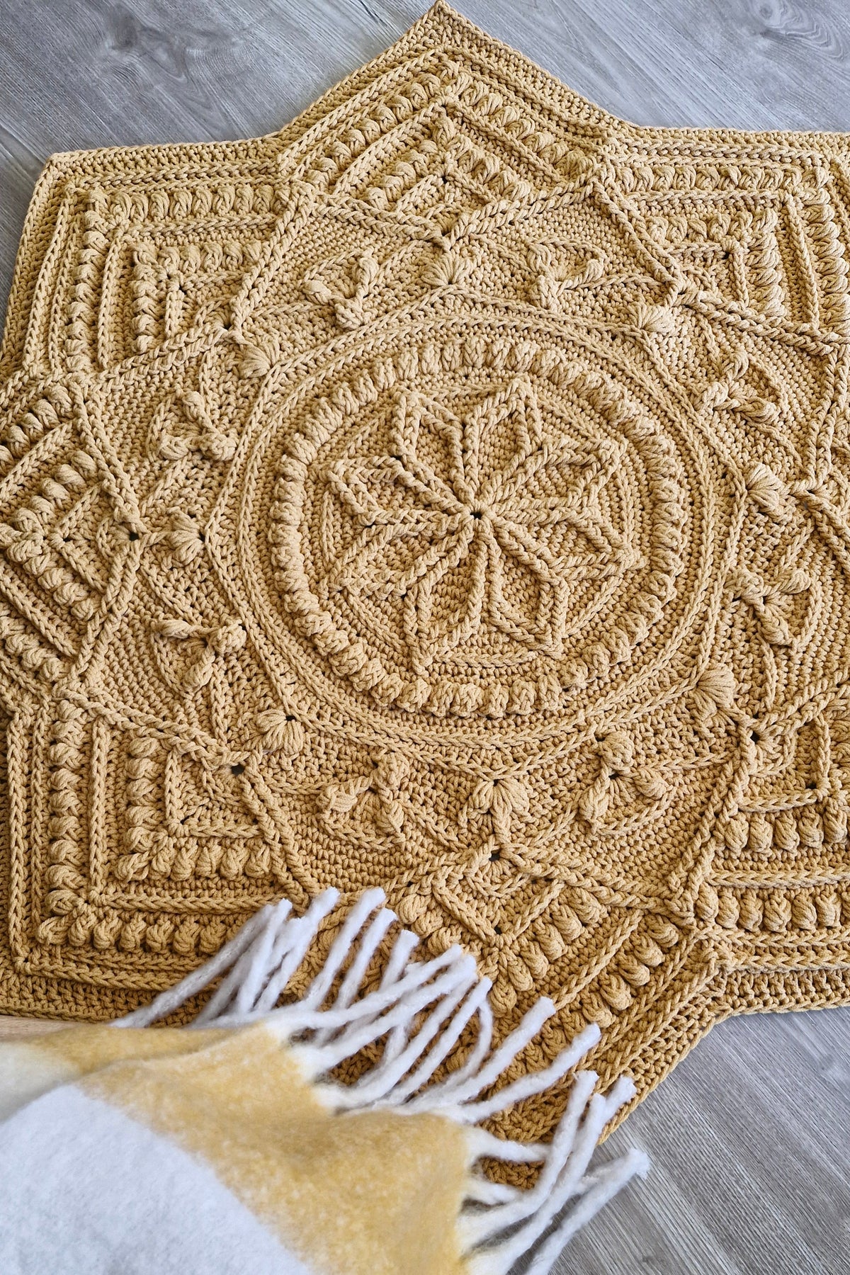 A large crocheted star-shaped crocher rug in a warm mustard-golden tan, laid flat on a gray wood floor. A white and mustard throw blanket with white fringe is partially visible in the lower foreground.