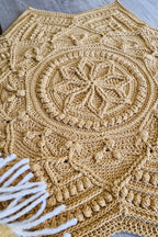 A handmade star-shaped crochet floor rug in a warm mustard color, featuring intricate circular and geometric patterns, styled on a gray wood floor with white fringe yarn visible in the lower left corner.