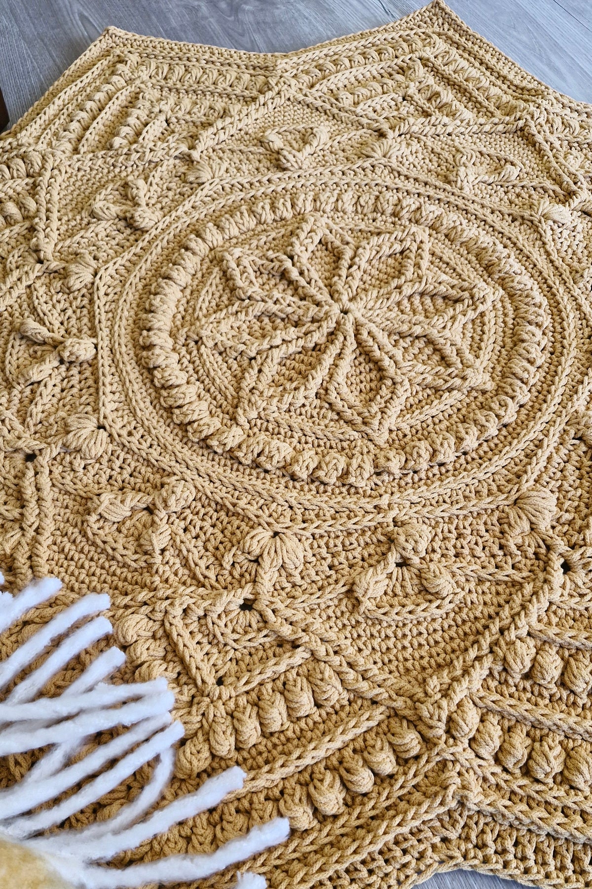 A handmade star-shaped crochet floor rug in a warm mustard color, featuring intricate circular and geometric patterns, styled on a gray wood floor with white fringe yarn visible in the lower left corner.