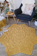 A large star-shaped crocheted area rug in mustard yellow is styled in a cozy living room setting on a light wood floor, next to a dark blue armchair with a floral pillow and a small wooden side table.