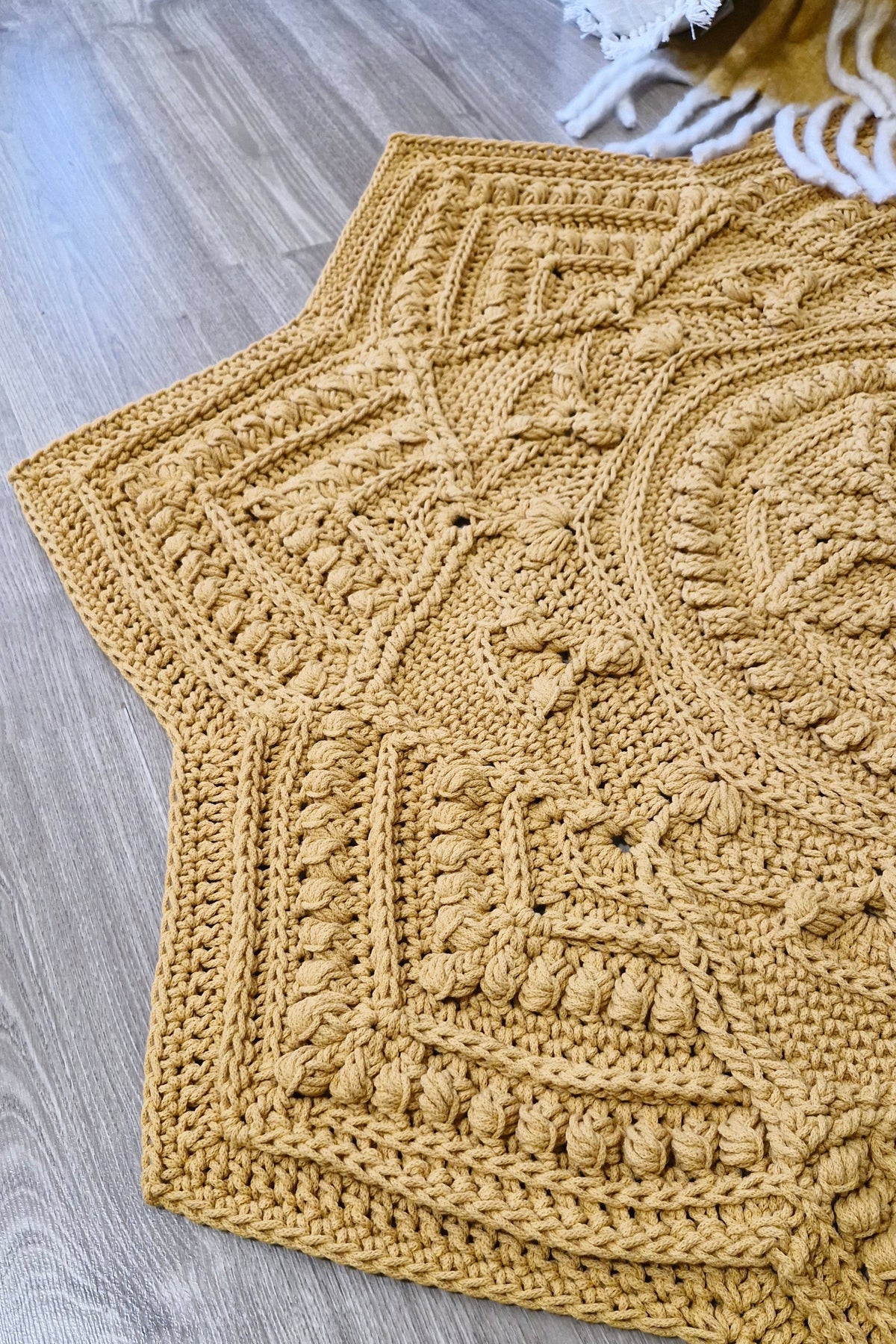A close-up of a chunky crocheted floor rug in a warm mustard yellow color, laid out on a gray wood floor.