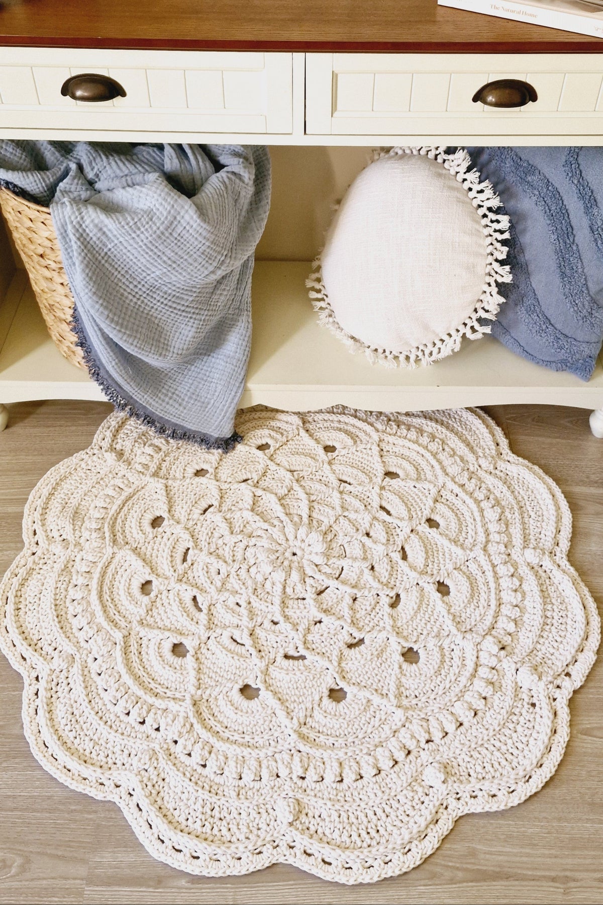 A round, off-white crocheted doily rug with an intricate floral mandala pattern, styled on a light wood floor in front of a white console table with a wooden top.