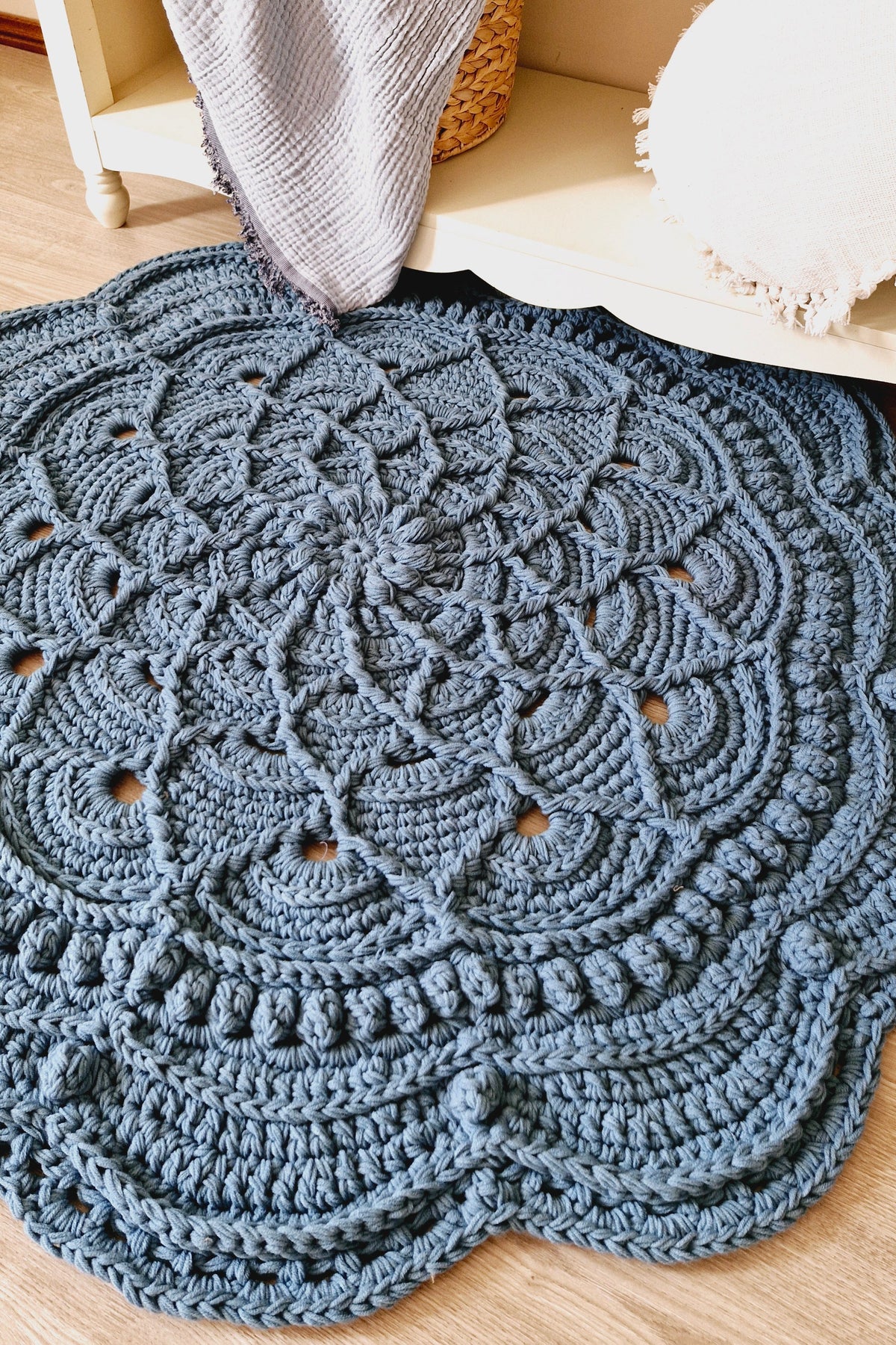 A round, chunky crocheted rug in a muted slate blue-gray color, styled on a light wood floor next to a white shelf with a textured blanket and a white pillow.