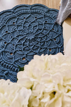 A dark blue crocheted doily floor rug with an intricate scalloped and floral lace pattern, laid out on a light wood floor next to a white fabric and a blurred foreground of white flowers.