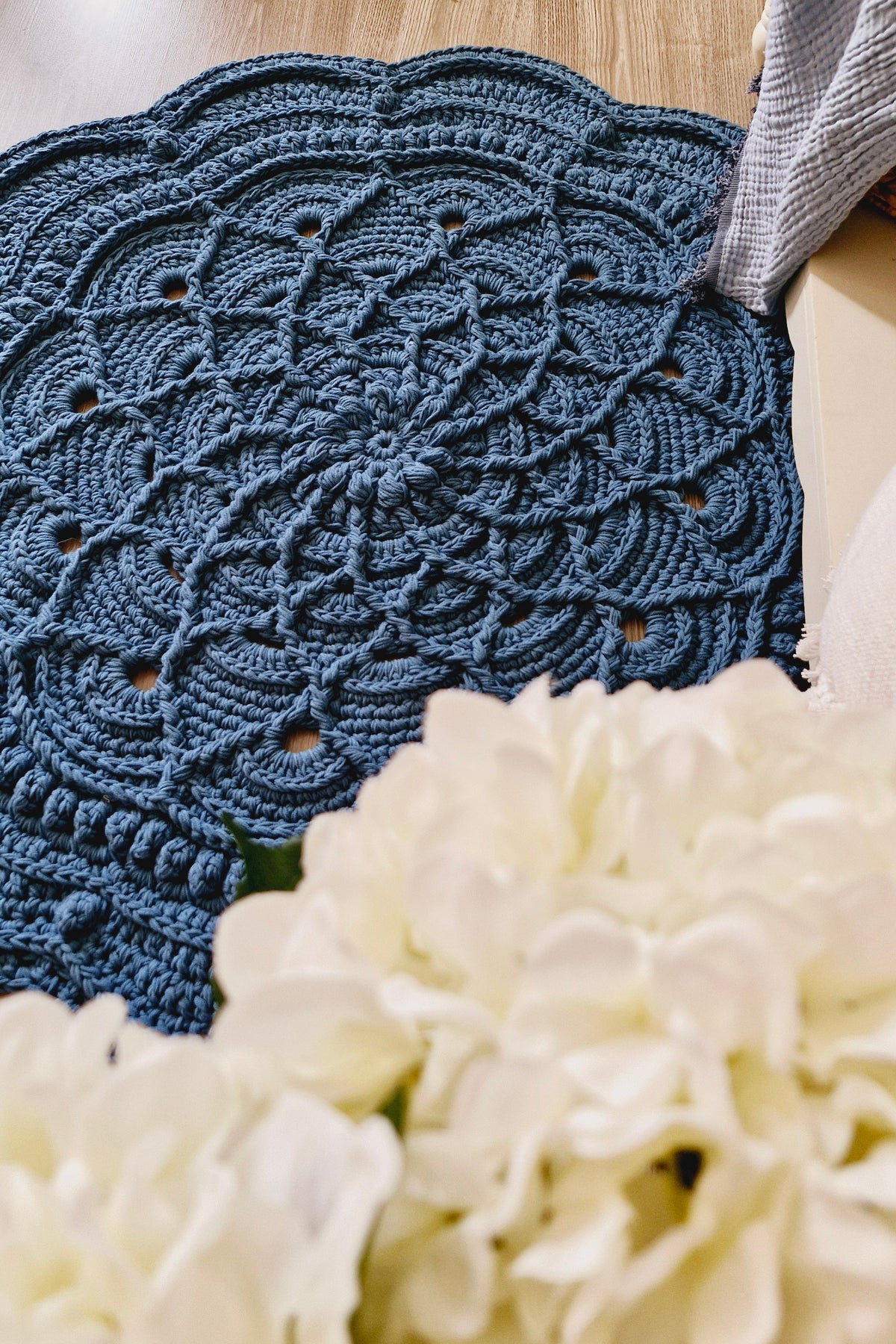 A dark blue crocheted doily floor rug with an intricate scalloped and floral lace pattern, laid out on a light wood floor next to a white fabric and a blurred foreground of white flowers.