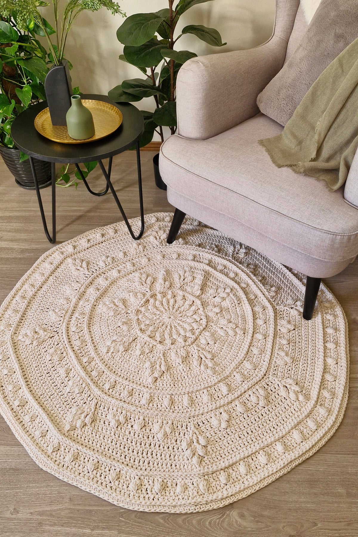 A round hand-crocheted area rug in a warm cream color, styled in a cozy living room setting next to a light beige armchair and a small black side table with plants.