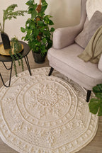 A round, cream-colored crocheted area rug with an intricate mandala and floral pattern, styled in a cozy living room setting next to a light gray armchair and potted plants.