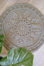 A round hand-crocheted rug with an intricate floral and leaf mandala pattern, styled on a light wood floor with green monstera leaves in the foreground.