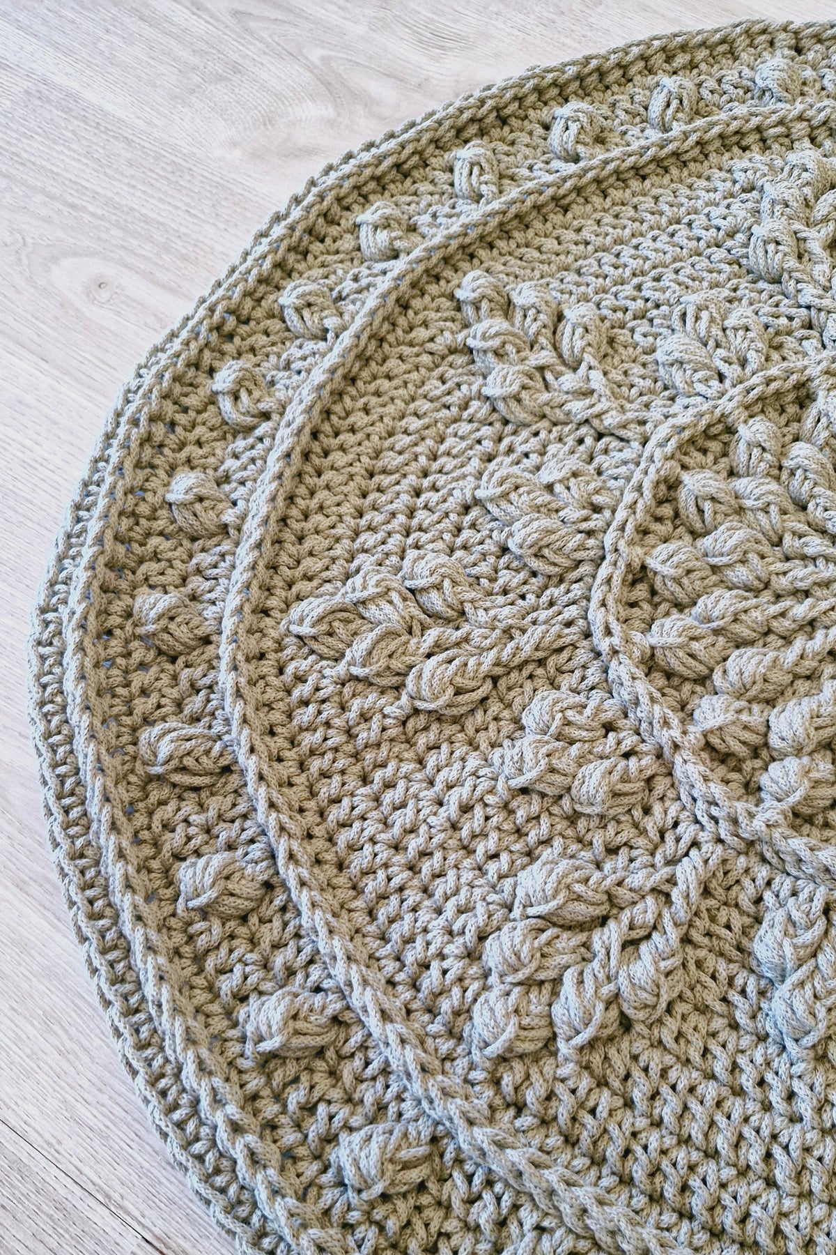 A close-up of a round, hand-crocheted rug in a green color, laid out on a light-colored wooden floor. The rug features a textured, textured crochet pattern arranged in concentric circular.