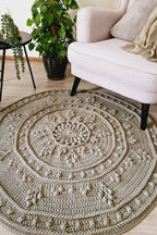 A round, hand-crocheted area rug in a warm green color, styled in a cozy living room setting next to an armchair and a potted green plant.