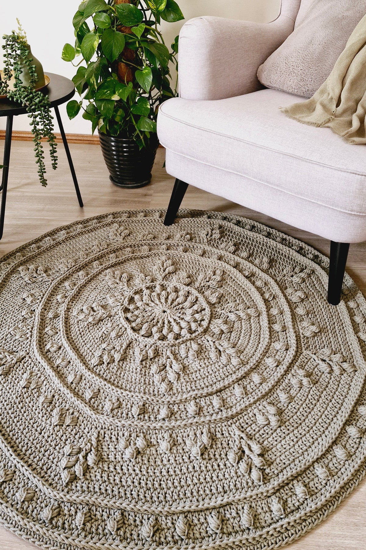 A round, hand-crocheted area rug in a warm green color, styled in a cozy living room setting next to an armchair and a potted green plant.