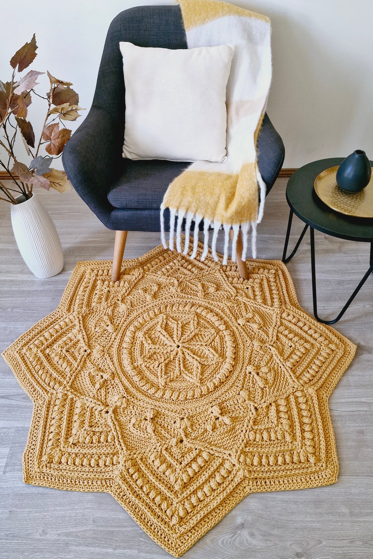 A large, intricate hand-crocheted area rug in a warm mustard yellow color, styled in a modern living room setting on a light wood floor next to a dark gray armchair and a small black side table.