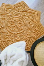 A large crocheted star-shaped textile piece in a warm mustard yellow is laid out on a light wood floor. A white plush throw blanket and a black bowl are partially visible in the foreground.