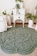 A large round crocheted rug in a muted sage green color, styled on a light wood floor in a cozy room setting with a white vintage nightstand, potted plants, and a white pillow.
