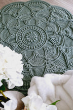 A round, sage green crocheted placemat with an intricate mandala pattern, styled on a wooden surface with white flowers and a white throw blanket.