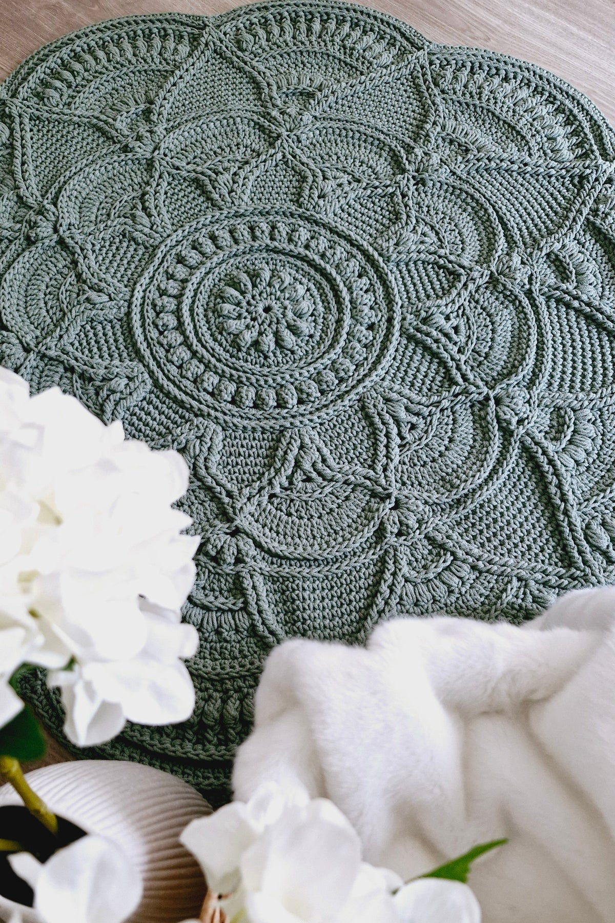 A round, sage green crocheted placemat with an intricate mandala pattern, styled on a wooden surface with white flowers and a white throw blanket.