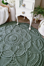 A round, hand-crocheted area rug in a muted sage green color, styled in a cozy room setting next to a white vintage nightstand and potted plants.