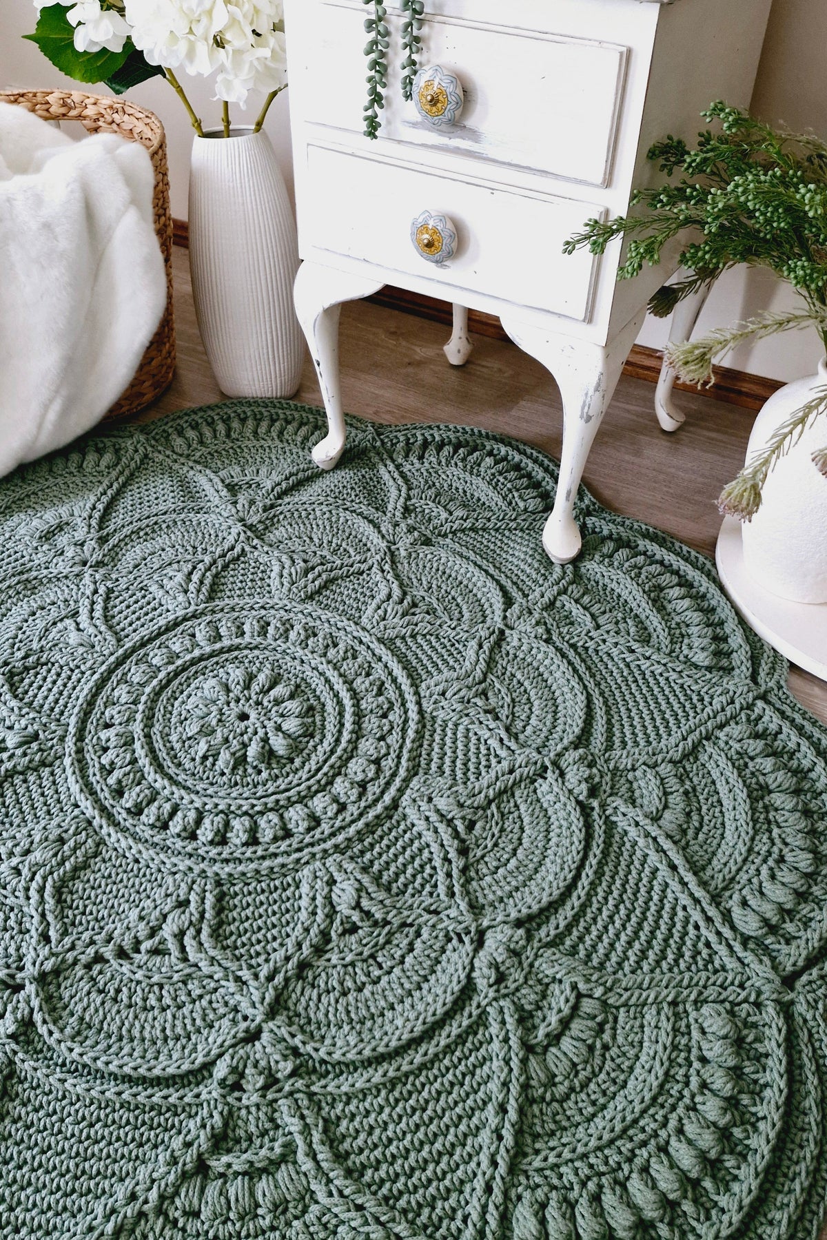 A round, hand-crocheted area rug in a muted sage green color, styled in a cozy room setting next to a white vintage nightstand and potted plants.
