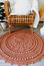 A round, hand-crocheted rug in a terracotta rust color, styled on a gray wood floor in front of a rattan armchair with a white throw blanket.