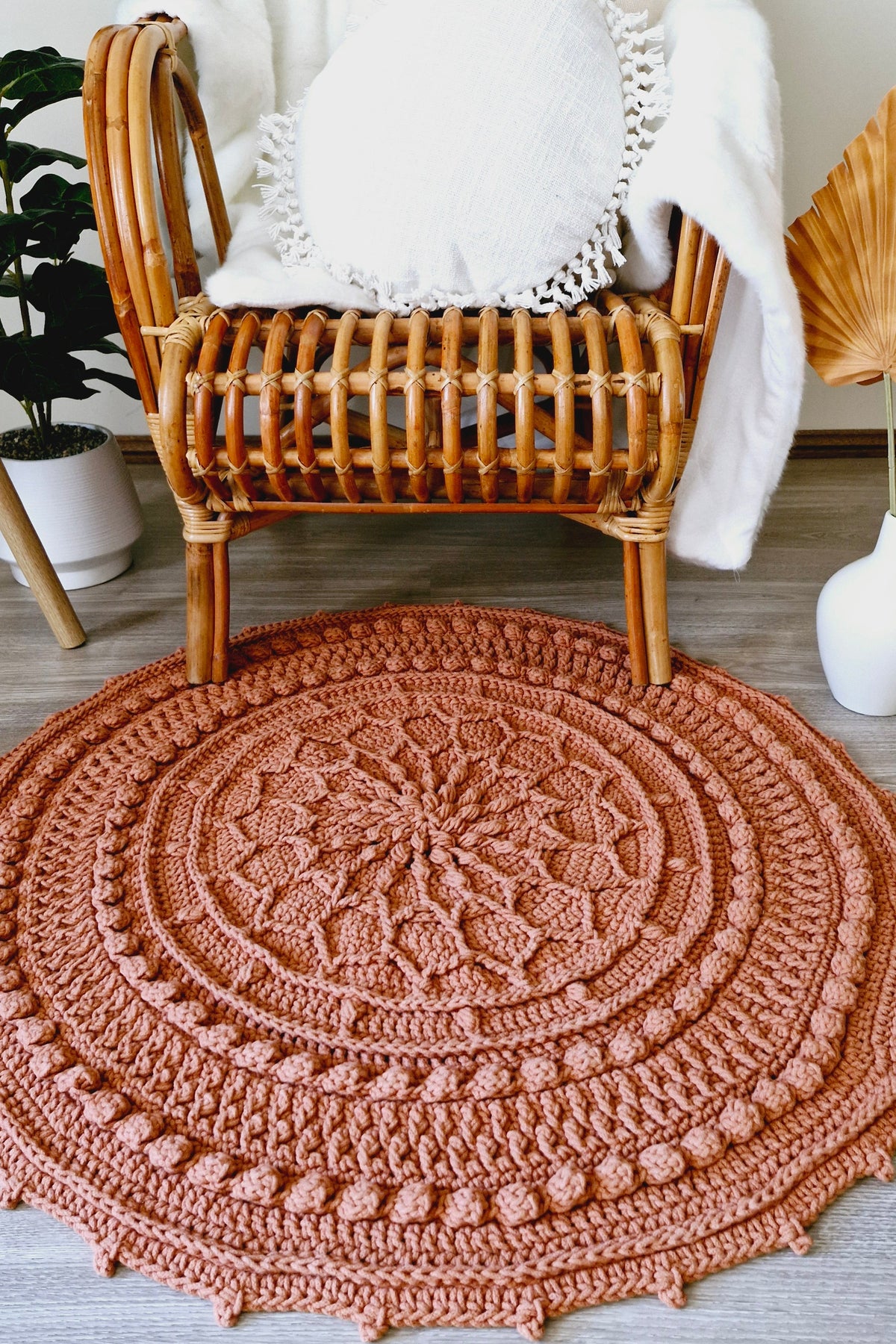 A round, hand-crocheted rug in a terracotta rust color, styled on a gray wood floor in front of a rattan armchair with a white throw blanket.