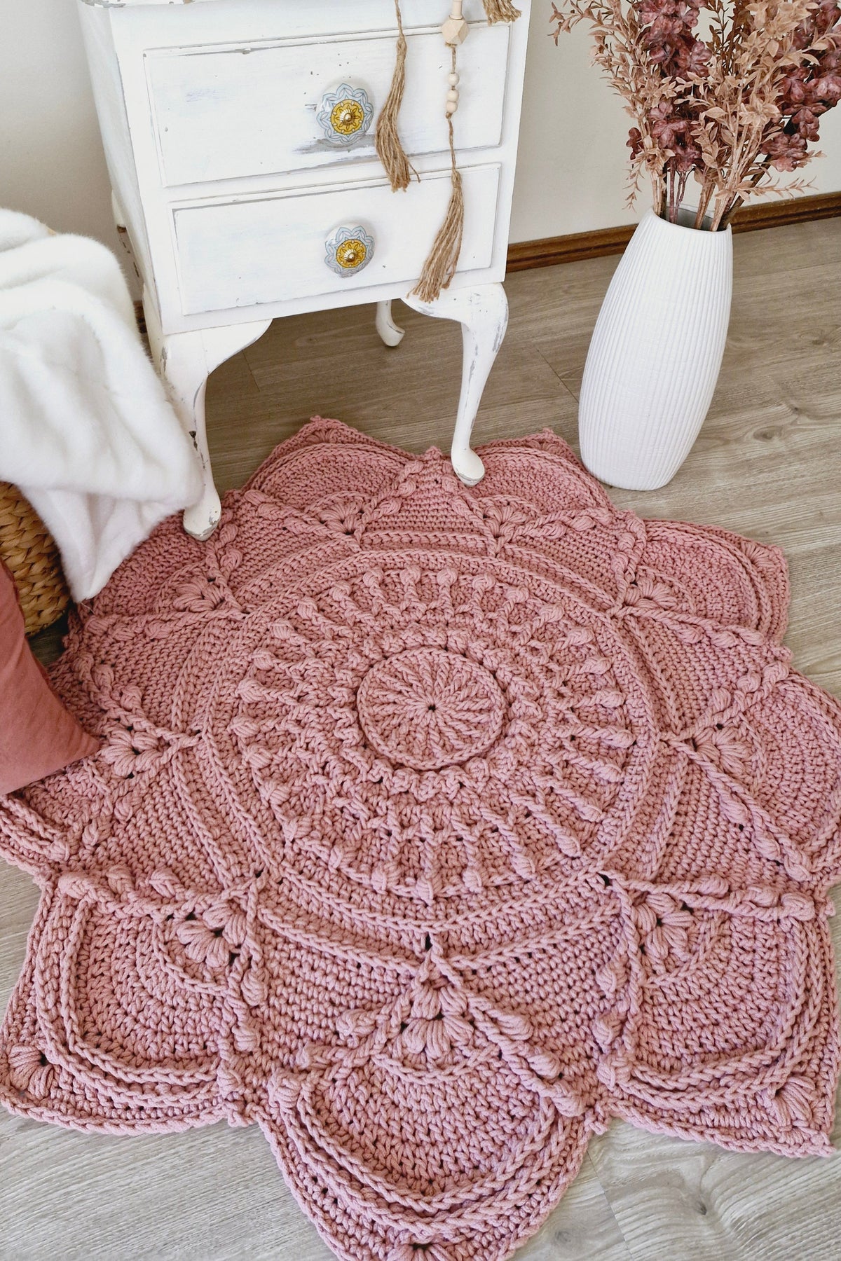 A large, round crocheted rug with intricate mandala and floral patterns in a dusty rose pink color, styled on a light wood floor next to a white vintage nightstand and a white vase with dried flowers.
