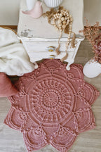 A round, hand-crocheted pink rug with an intricate mandala pattern, styled on a light wood floor next to a distressed white nightstand with decorative items.