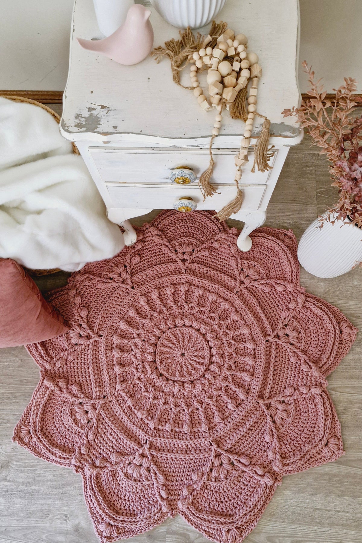A round, hand-crocheted pink rug with an intricate mandala pattern, styled on a light wood floor next to a distressed white nightstand with decorative items.
