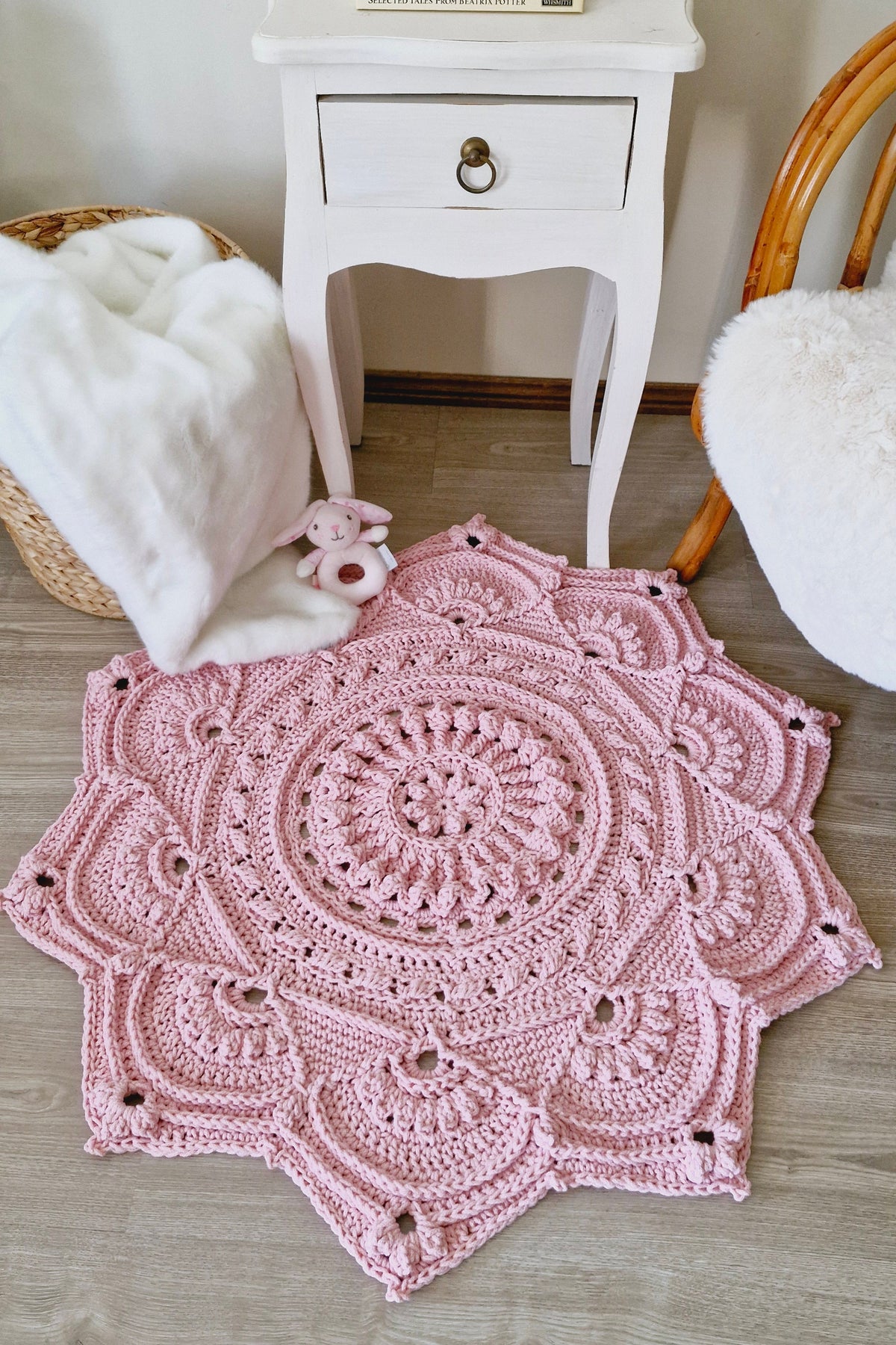 A pink crocheted floral-shaped rug with an intricate mandala pattern, styled on a light wood floor in a cozy room setting with a white side table, a woven basket, and a wooden chair.