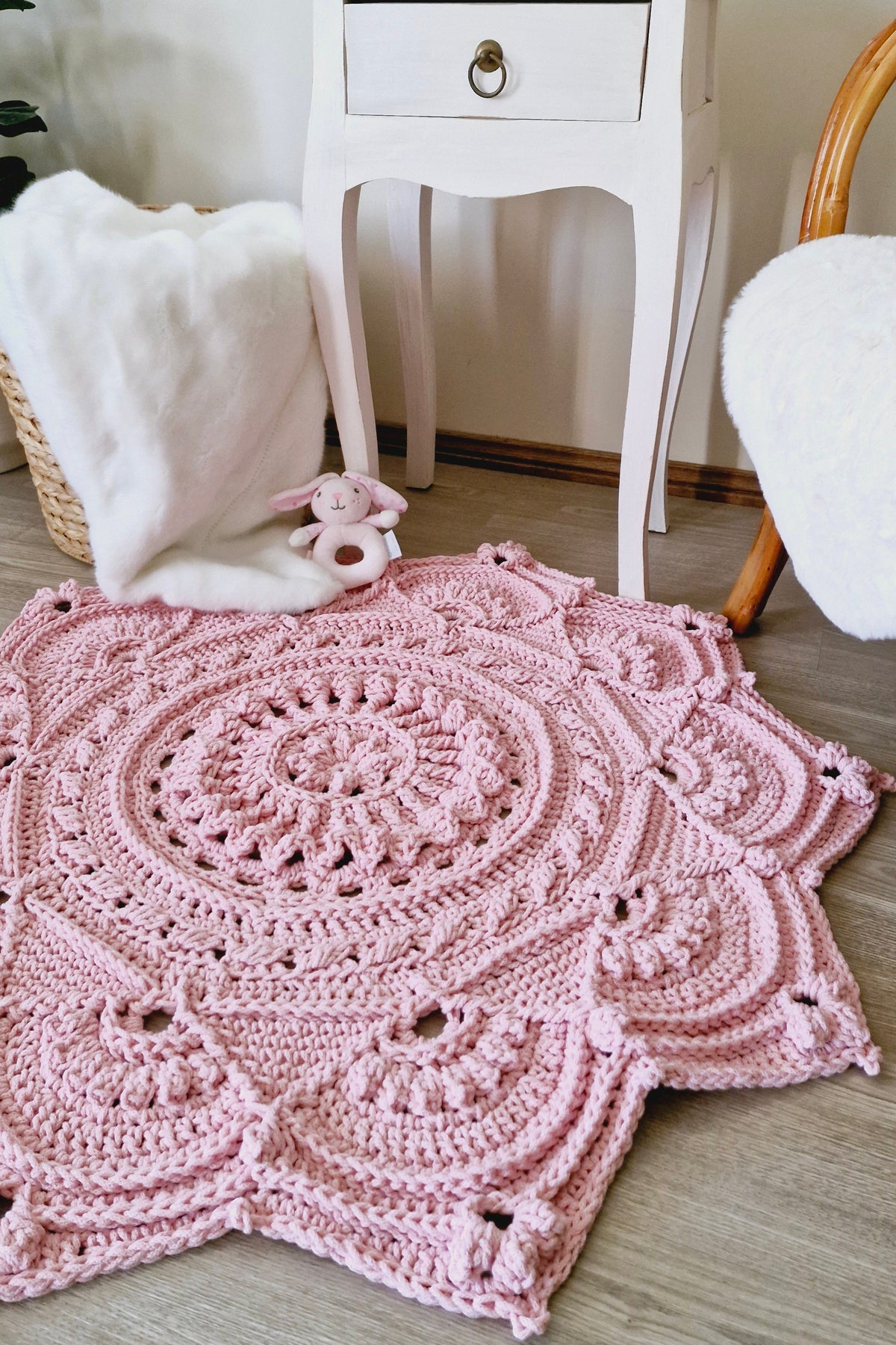 A pink crocheted floral-shaped rug with an intricate mandala pattern and scalloped edges, styled on a light wood floor next to a white side table and a white plush throw blanket.