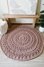 A round, hand-crocheted rug in a dusty brown color, styled on a light wood floor in a cozy room setting with a wooden bench, framed wall art, and a potted plant.
