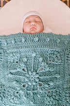 A sleeping newborn baby wearing a white hat, nestled under a textured, light blue-green crocheted blanket with a floral pattern, lying on a white circular mattress.