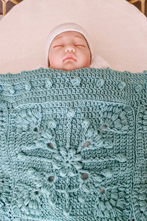 A sleeping newborn baby wearing a white hat, nestled under a textured, light blue-green crocheted blanket with a floral pattern, lying on a white circular mattress.